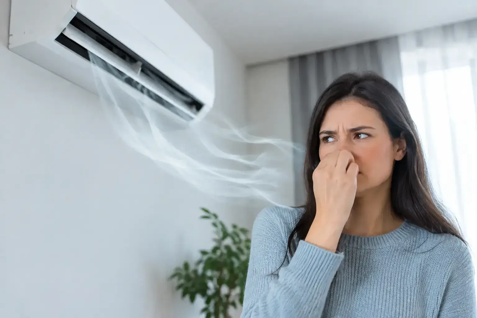 Woman in a grey sweater pinching her nose and frowning at a wall-mounted ductless air conditioner as wisps of air blow out of it